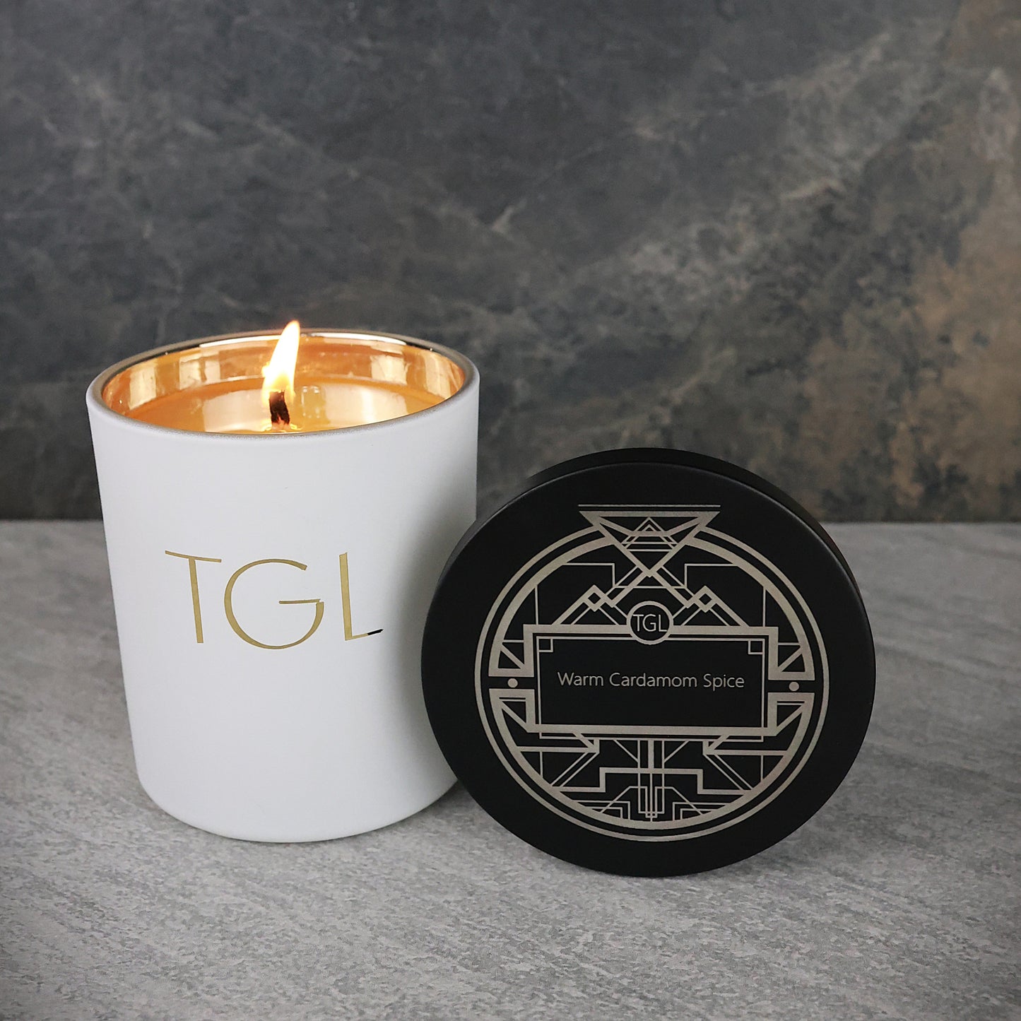 White candle with gold interior and 'TGL' branding next to a black coaster on a gray surface.