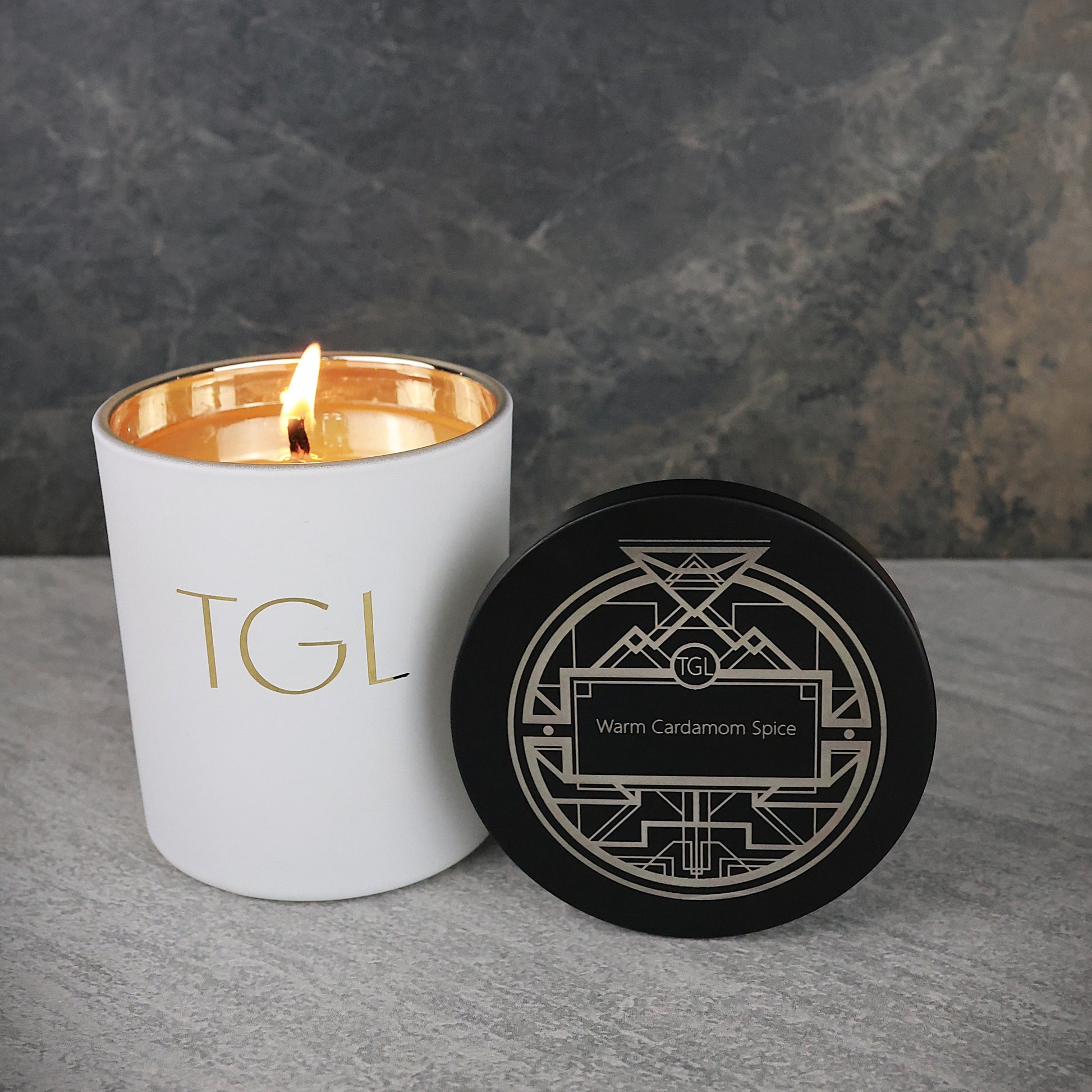 White candle with gold interior and 'TGL' branding next to a black coaster on a gray surface.