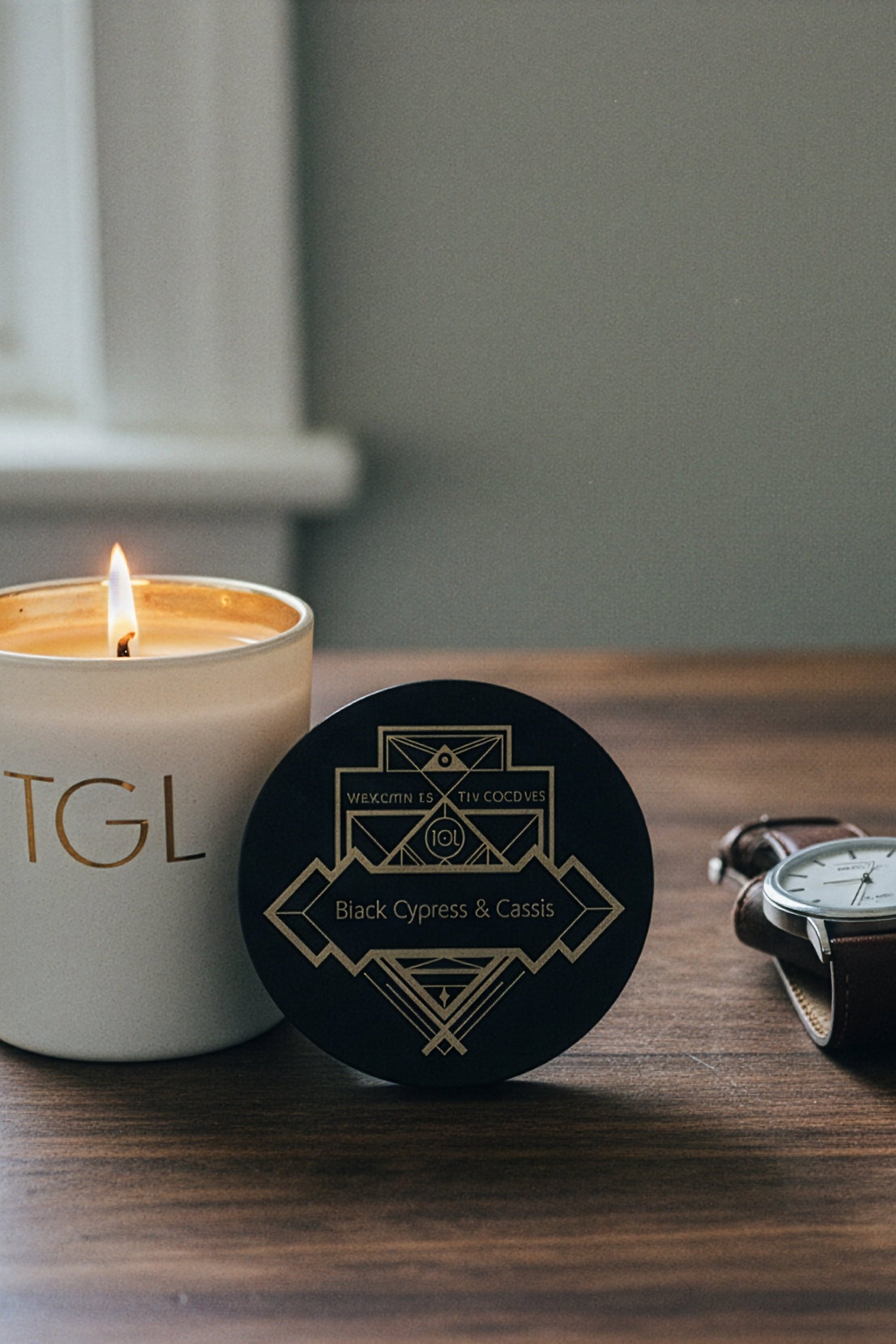 A lit white soy candle with gold lettering sits on a wooden table next to a round black lid with a gold geometric logo and the words 'The GOOD LIFE.' A dark leather wallet, a wristwatch, and a glass of amber liquor are arranged nearby in a moody indoor setting. Soy Candle, Soy beeswax Candle.