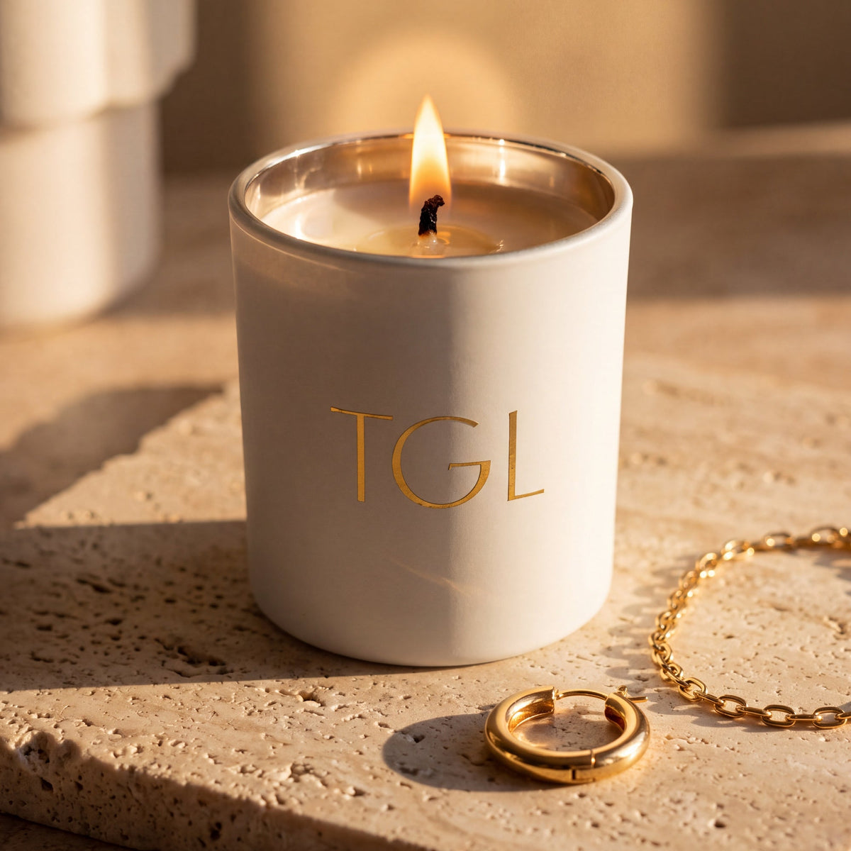 TGL soy candle by The GOOD LIFE, lit in a white jar in warm sunlight beside a gold bracelet