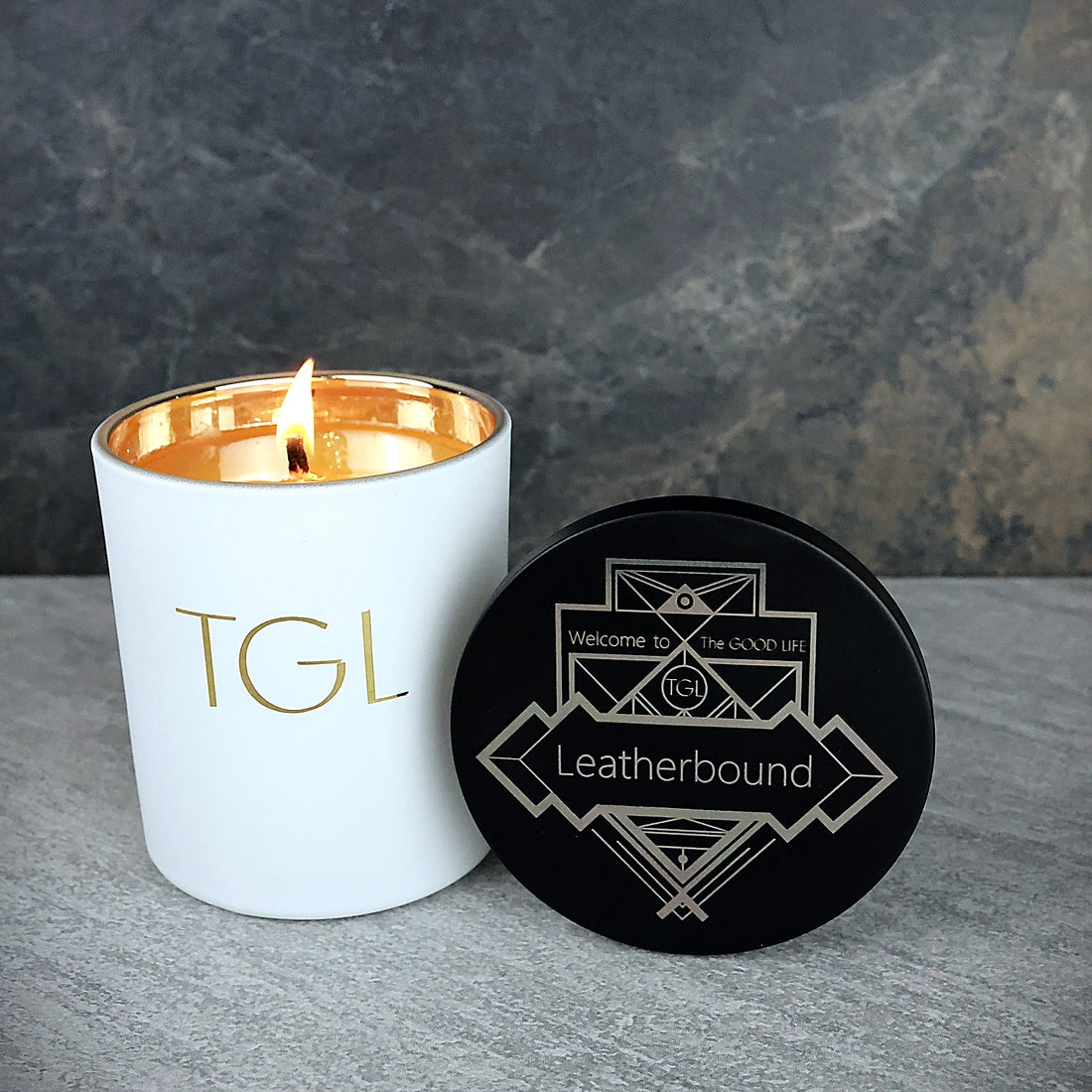 Lit Leatherbound TGL soy candle beside its black Art Deco engraved lid